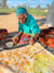 Artist Polly Ngale wears a turquoise polo and matching headscarf. She is outside, kneeling on a large blanket and is stretching over a painting with a brush in her hand, facing towards the camera. It is a sunny day. She is painting a daub of white paint.